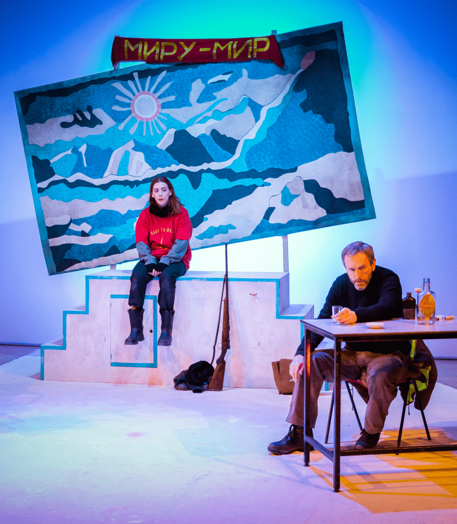 Performer Rebecca Wilkie sits atop a podium at the back of the stage with her legs hanging off the edge. She wears a bright red tshirt with black trousers and boots. She looks downstage at performer Matthew Zajac who is sitting at a table drinking a glass of whiskey. They both look frustrated. In the background is a large blue tapestry of mountains and a sun, hung squint and a red fabric banner with Russian text in yellow letters.
