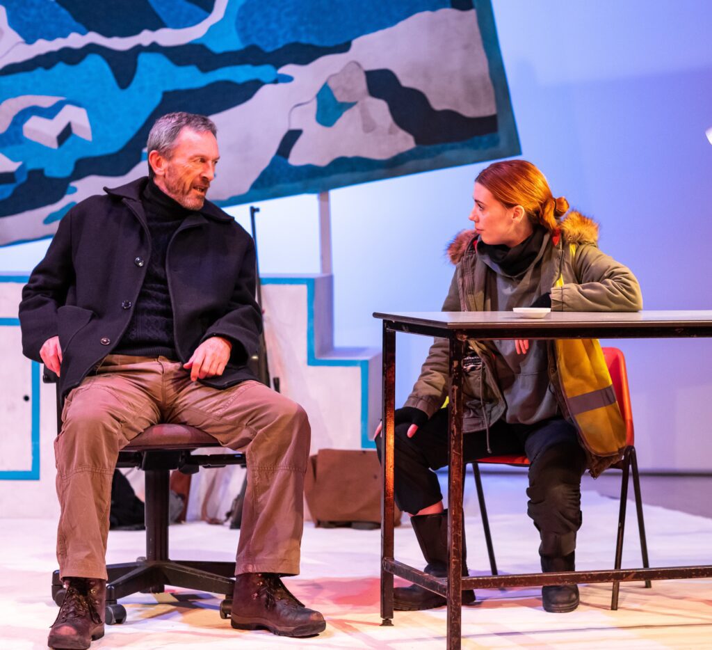 Performers Matthew Zajac and Rebecca Wilkie sit on chairs centre stage in earnest conversation. Matthew wears grey trousers and a heavy grey coat. Rebecca wears a khaki parka with a bright neon vest over the top and big black boots. Behind them is a large blue tapestry of mountains and a sun, hung squint.