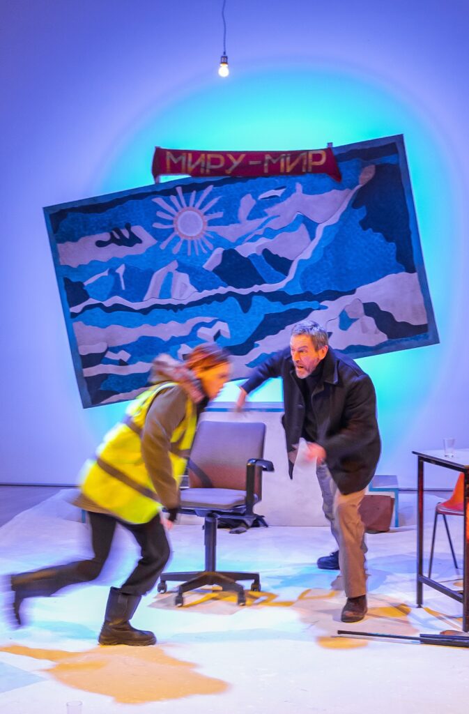 Performer Rebecca Wilkie runs to the right whilst performer Matthew Zajac moves to block her. Matthew wears grey trousers and a heavy grey coat. Rebecca wears a khaki parka with a bright neon vest over the top and big black boots. In the background is a large blue tapestry of mountains and a sun, hung squint and a red fabric banner with Russian text in yellow letters.