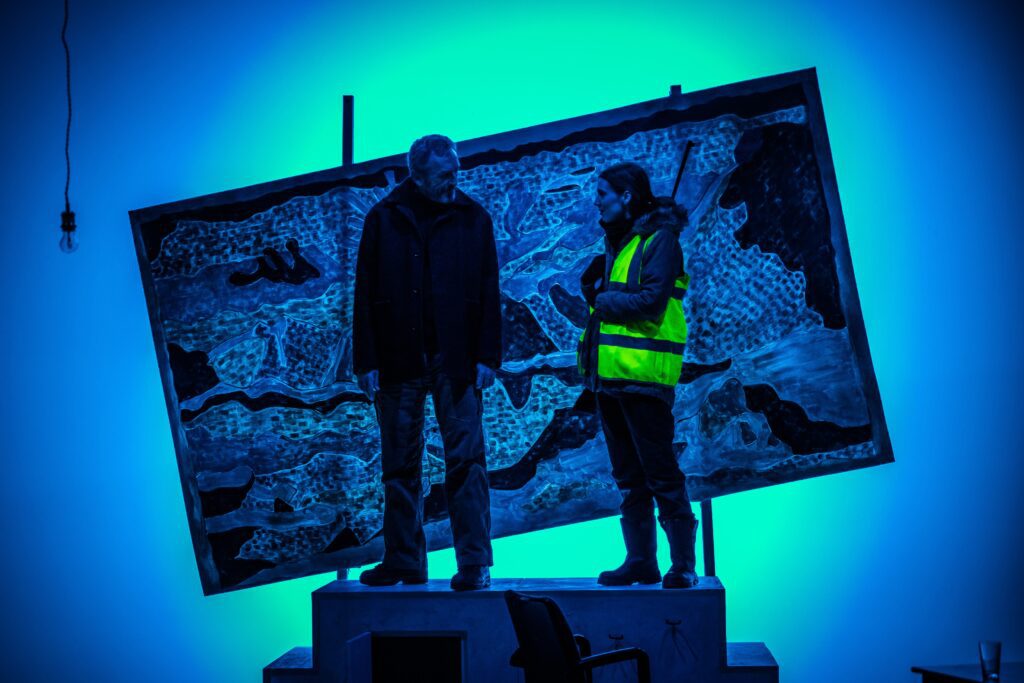 Still from Nightlands. Performers Matthew Zajac and Rebecca Wilkie stand atop a podium in conversation with one another. Matthew wears grey trousers and a heavy grey coat. Rebecca wears a khaki parka with a bright neon vest over the top and big black boots. Behind them is a large blue tapestry of mountains and a sun, hung squint.