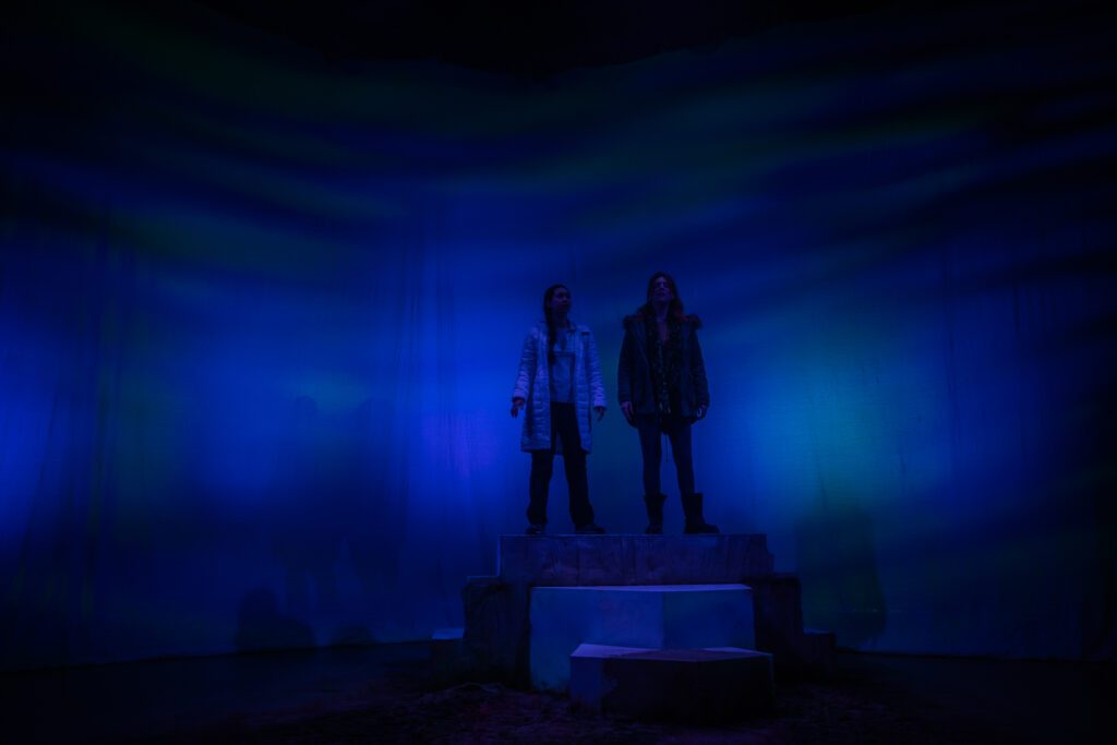 Against a dimly lit blue backdrop, performers Rebecca Wilkie and Tara McGirr stand atop a block and look out to the sky. Rebecca wears a khaki parka jacket with a fur hood, a green scarf, skinny jeans and chunky black boots. Tara wears a knee length white puffer jacket with her hair in long plaits.