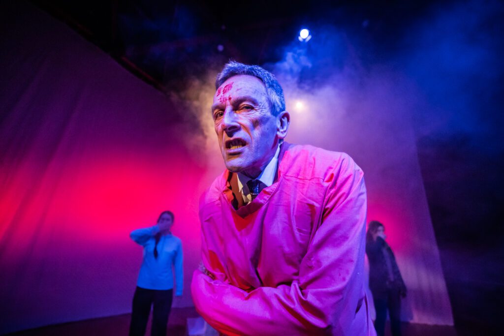 Performer Matthew Zajac stares into the camera with blood across his forehead and cheek. He wears a red straight jacket over his brown suit. Behind him, performers Rebecca Wilkie and Tara McGirr cover their mouths in from of a red backdrop.