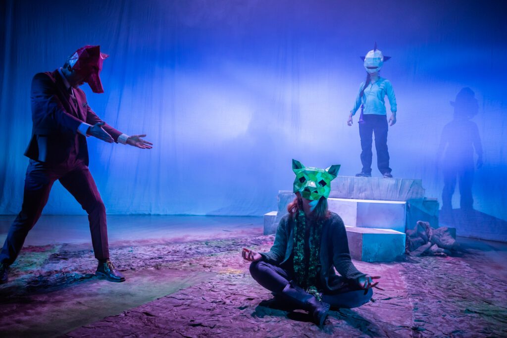 Still from Fallen Angels of the Moine by George Gunn. In the centre, performer Rebecca Wilkie sits cross-legged on the floor wearing a green pig mask. Performer Matthew Zajac stands to her left wearing a red fox mask and a brown suit, with his arms out towards her. Tara McGirr stands on a podium in the background wearing a blue whale mask and looking down at Rebecca.