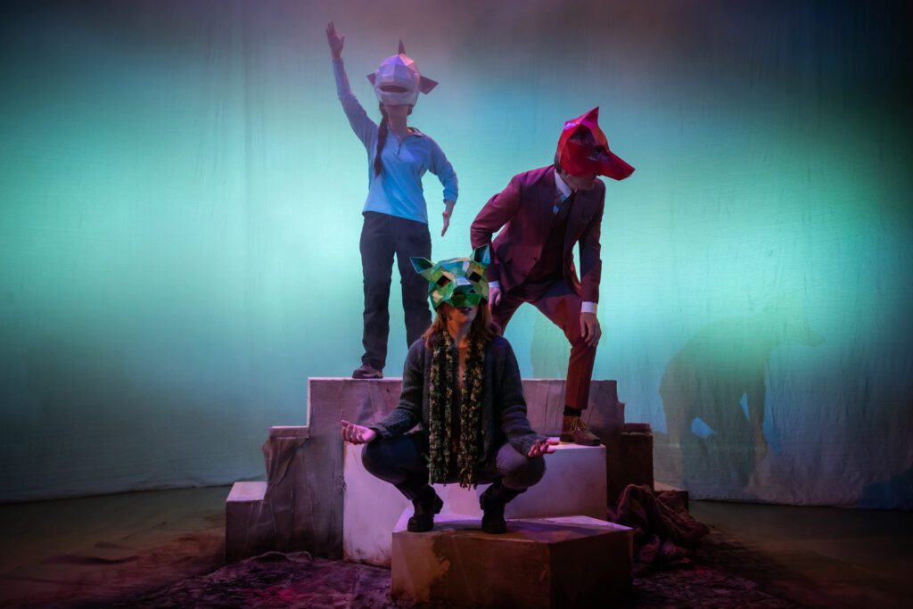 Still from The Fallen Angels of the Moine by George Gunn. Three performers, Matthew Zajac, Rebecca Wilkie and Tara McGirr, pose in front of a blue-washed backdrop. Rebecca crouches at the front wearing a green pig mask with her hands resting on her knees. Matthew leans to the right wearing a red fox mask. Tara stands at the back wearing a blue whale mask, reaching one hand up to the sky. Company
