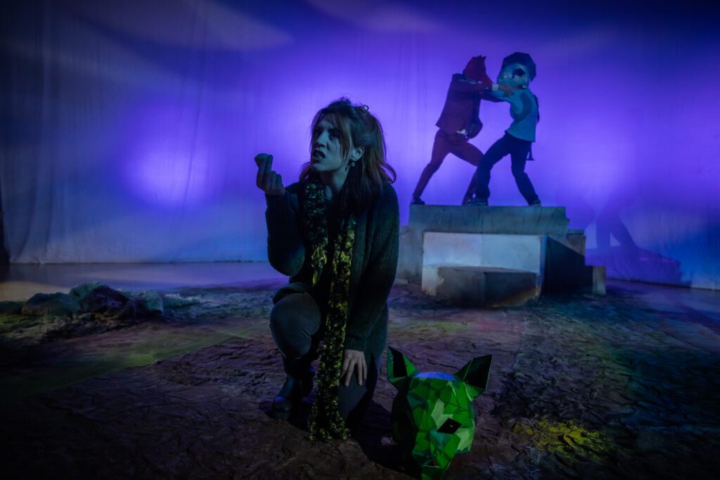 Performer Rebecca Wilkie crouches at the forefront next to a green pig mask, holding a stone up in one hand. In the background, a performer with a red fox mask and a performer with a blue whale mask wrestle with one another on a podium. The backdrop is purple lit cloth and the floor of the space is brown and green, reminiscent of dirt.