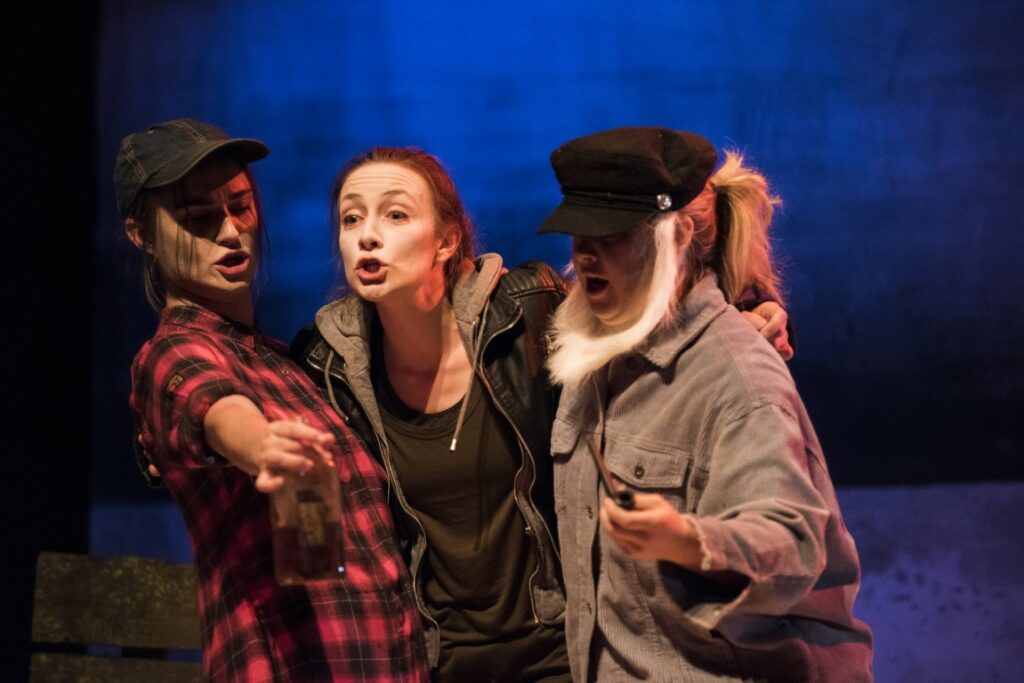 Still from The Stornoway Way. Performer Naomi Stirrat lurches forward shouting with her arms wrapped around two other performers, Chloe-Ann Taylor on the left and Rachel Kennedy on the right. Naomi wears a black tshirt with a grey hoodie and black leather jacket. Chloe-Ann wears a red plaid shirt with a black baseball cap and holds out a bottle of whiskey. Rachel wears a faux white beard with a black paperboy cap and grey corduroy shirt. She holds a black smoking pipe.