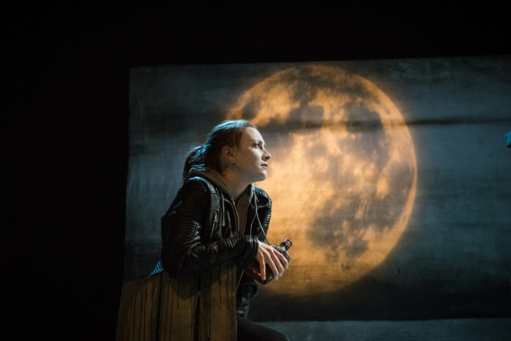 Performer Naomi Stirrat looks wistfully out to the right, holding a bottle of whiskey. She wears a grey hoodie under a leather jacket. She leans up against a fence in front of a backdrop of a large yellow moon.