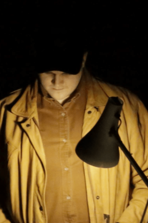 About Us – Jack MacGregor A portrait of Jack MacGregor whilst he performs and extract of his work. Jack is wearing a mustard shirt and brown canvas jacket with a black baseball cap. He looks down at a script.